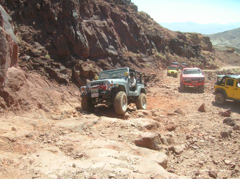 1985 Ford Bronco - Multiple off-road vehicles traverse a challenging rocky path in a desert landscape.