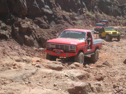 1985 Ford Bronco - A convoy of off-road vehicles navigates a , rocky trail under a bright sun.