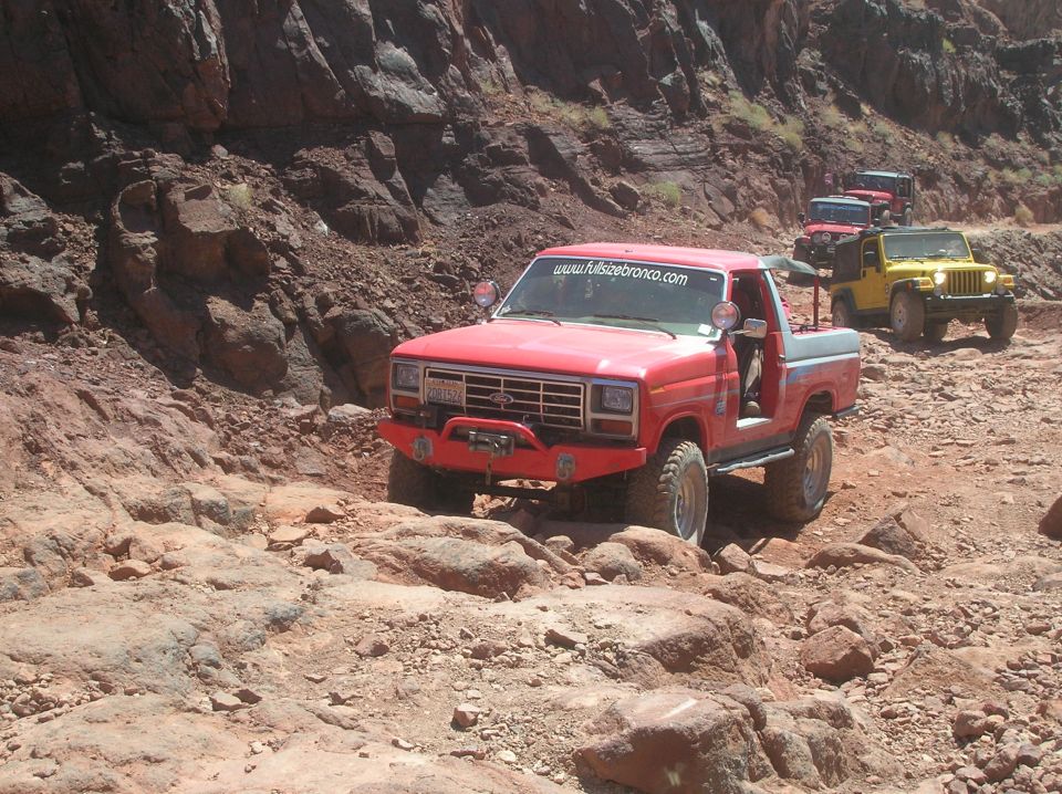 red pickup truck with "www.fullsizebronco.com" on windshield driving on rocky terrain, followed by other off-road vehicles