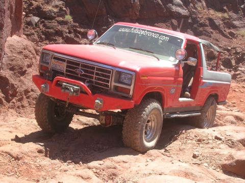 1985 Ford Bronco - A red Ford Bronco navigates a challenging rocky terrain, its front end angled upwards.