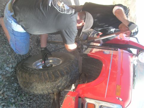 1985 Ford Bronco - Two people are seen working on the front wheel of a red vehicle, with one person using a jack.