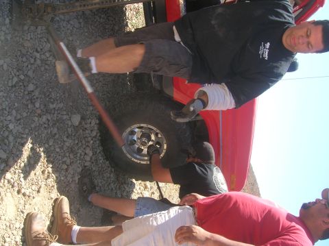 1985 Ford Bronco - Three men work together to change a flat tire on a red SUV in a dry, outdoor environment.
