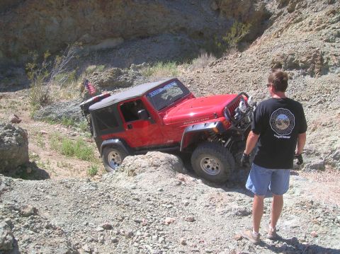 1985 Ford Bronco - A red Jeep Wrangler Rubicon tackles a challenging rocky terrain, with an American flag visible on the rear.
