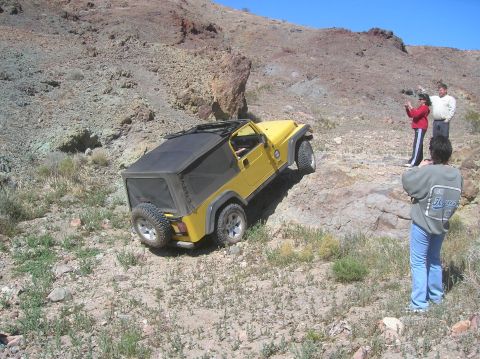 1985 Ford Bronco - A yellow Jeep Wrangler navigates a , rocky terrain under a clear blue sky.
