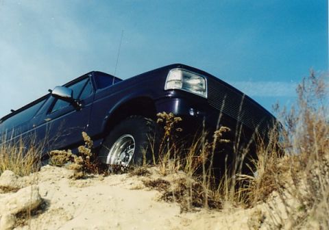 1994 Ford Bronco - A dark blue SUV is partially obscured by dry grass and sand, viewed from a low angle against a clear blue sky.