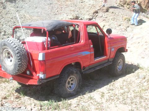 1985 Ford Bronco - This red off-road SUV, likely from the early 1980s, is seen on a rocky terrain with people in the background.