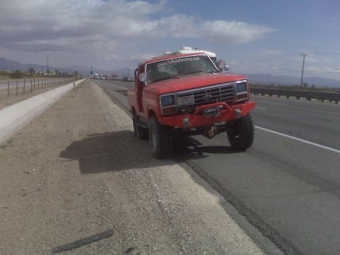 1985 Ford Bronco - A red Ford Bronco is parked on the side of a highway under a cloudy sky.