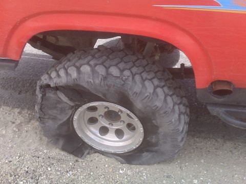 1985 Ford Bronco - A red SUV with a flat tire, likely a 1985 Ford Bronco, is seen with its sidewall severely damaged.