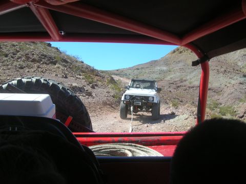 1985 Ford Bronco - A white off-road vehicle is seen on a dusty trail, with another vehicle's interior visible in the foreground.