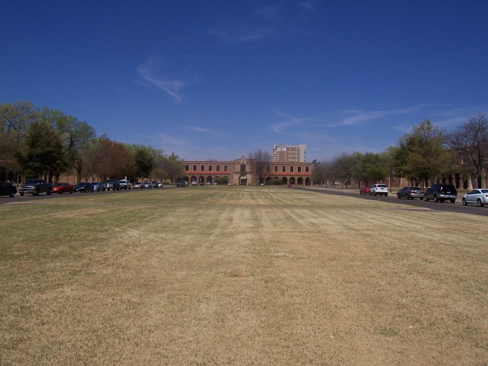 Cars parked along a road in front of a long, arched building under a clear blue sky