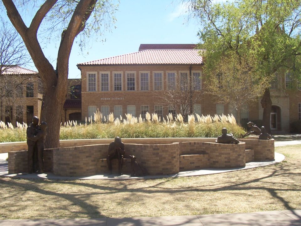 Building with "HUMAN SCIENCES" text, brick facade, and tiled roof, surrounded by grass, trees, and sculptures