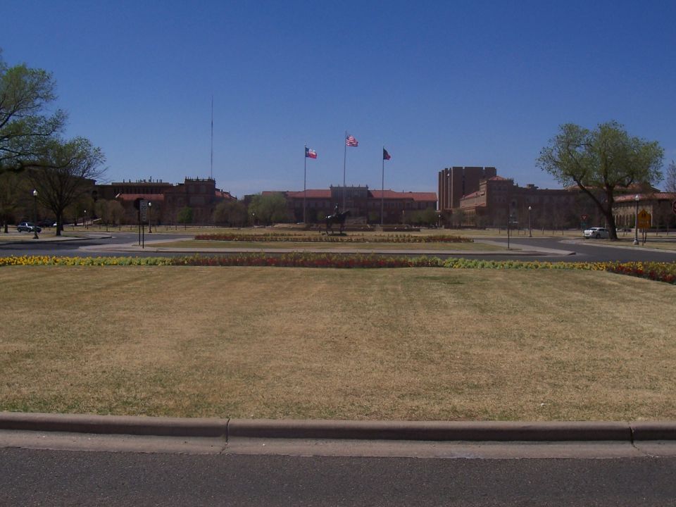 Campus scene with buildings, flags, and a statue under a clear blue sky