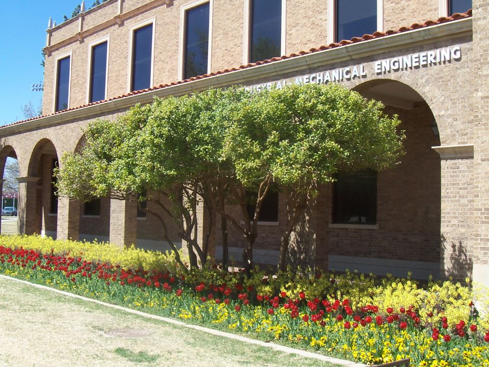 Building with arched walkways and windows, surrounded by trees and flower beds