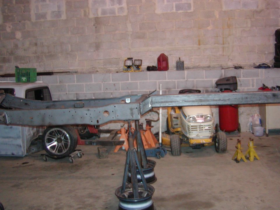 Vehicle frame supported by jack stands in a garage, with a lawn tractor and other equipment nearby