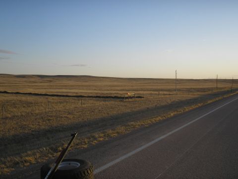 1990 Ford Bronco - A spare tire and jack are visible on the side of a dirt road, with a wide, open grassland stretching to the horizon under a clear sky.
