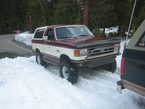 1995 Ford Bronco - A two-tone Ford Bronco sits partially buried in snow on a wooded road, with another vehicle visible to the right.