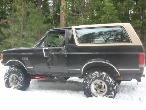 1995 Ford Bronco - A lifted black Ford Bronco with a tan top sits in the snow, showing damage to the driver's side door.