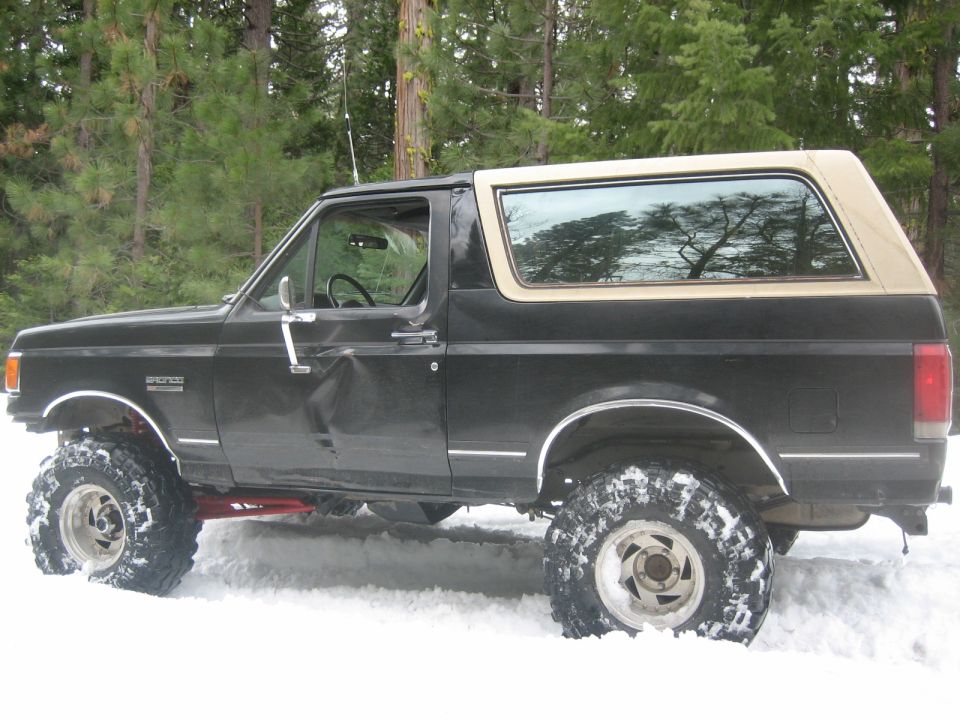 Black Ford Bronco with a tan removable top, lifted with large tires, in the snow. Dented driver's side door