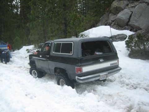 1995 Ford Bronco - A black Chevrolet K5 Blazer is stuck in deep snow on a wooded trail, with another vehicle visible in the background.