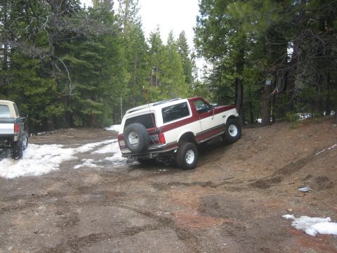 1995 Ford Bronco - Two SUVs navigate a snowy, wooded trail; the Bronco is on an incline.