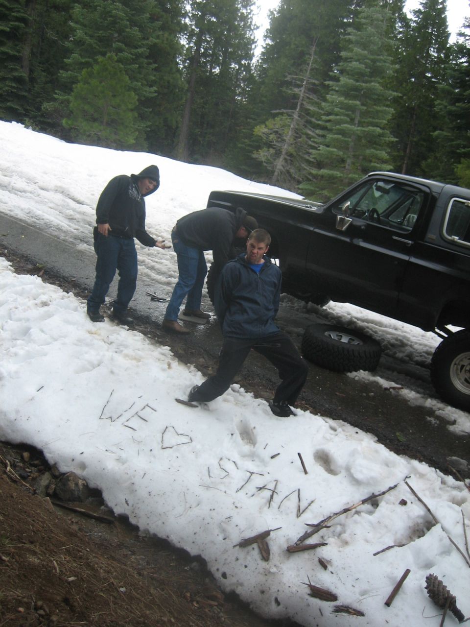 Black SUV with a removed tire in the snow, three people nearby. 'WE <3 STAN' written in snow