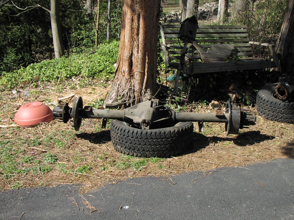 vehicle axle assembly sitting on a tire outdoors next to a tree and a wooden bench