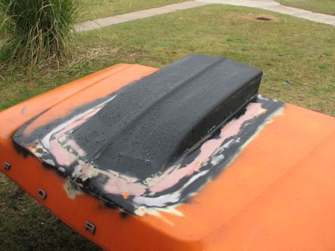 1979 Ford Bronco - Orange truck hood laid upside-down on a lawn, fitted with a raised black cowl-induction style hood scoop that has been rattle-canned black…