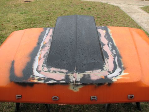 1979 Ford Bronco - An orange Ford hood with a raised center cowl/scoop section, viewed from the front while the hood rests on stands outdoors on grass. The ho…