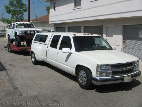 1994 Chevrolet Silverado 3500 - A white extended cab dually pickup truck tows a white SUV on a trailer, parked in front of a building.