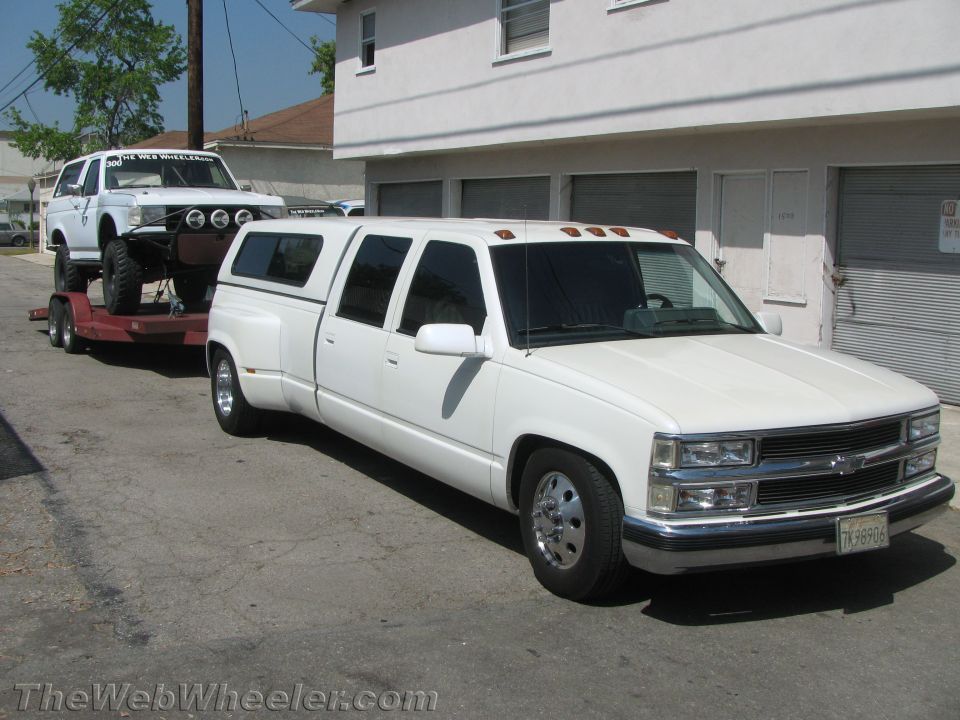 White extended cab dually pickup truck towing a white SUV on a trailer. Truck has a camper shell