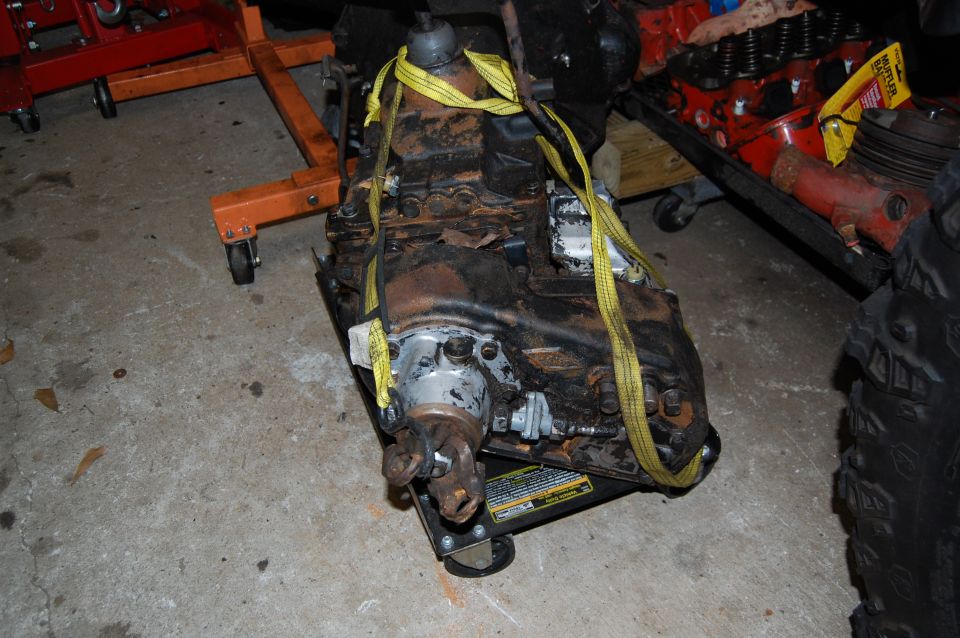 Transmission on a dolly, secured with yellow straps. Red engine parts and a tire are visible