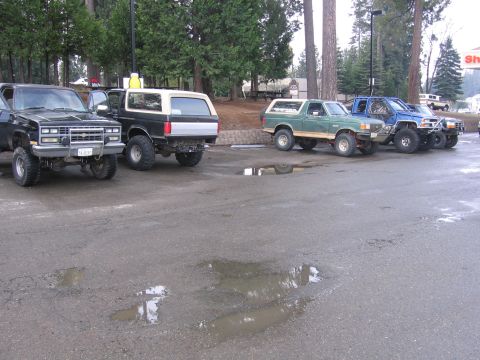 1989 Ford Bronco - Four off-road trucks are parked in a lot with puddles on the ground.