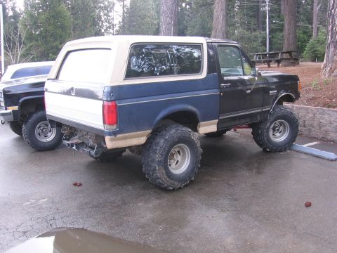 1989 Ford Bronco - A lifted blue and white Ford Bronco is parked next to another vehicle in a wooded area.