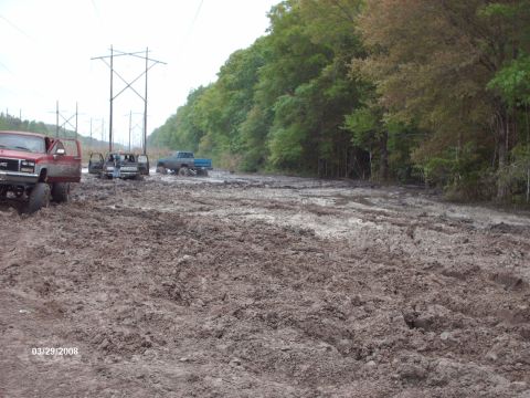 1982 Ford F-100 - Several trucks are stuck in deep mud on a wooded road, with power lines overhead.
