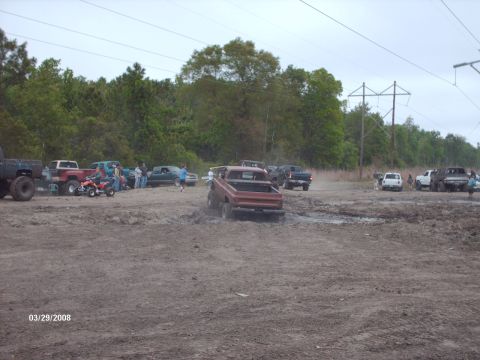 1982 Ford F-100 - Multiple trucks and ATVs are gathered in a muddy field, with one red pickup truck actively driving through the mire.