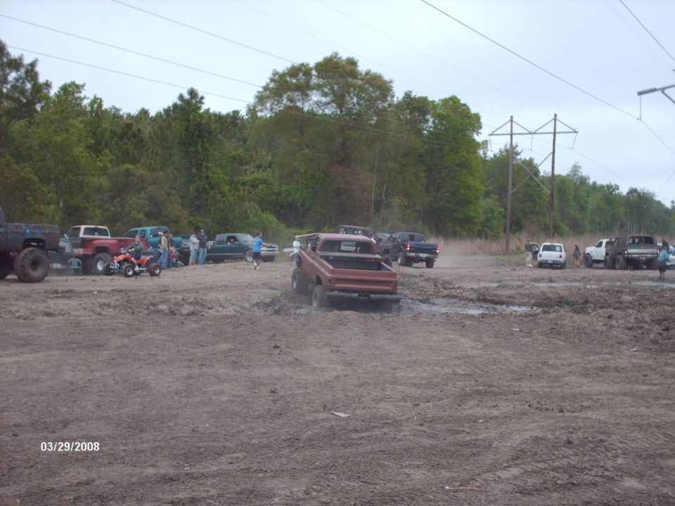 Red pickup truck with lifted suspension driving through mud. The truck has a stepside bed and appears to be from the early 1980s