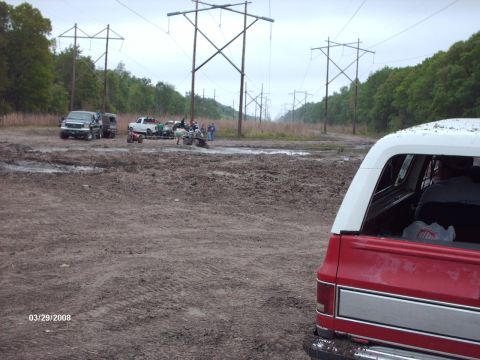 1982 Ford F-100 - Off-roading scene with multiple vehicles in a muddy field, with power lines stretching into the distance.