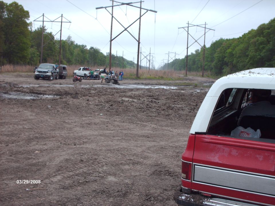 Muddy field with several trucks, ATVs, and power lines under an overcast sky. A red and white vehicle is in the foreground