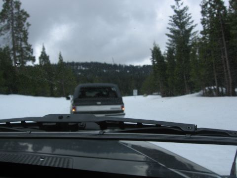 1989 Ford Bronco - A pickup truck is seen from inside another vehicle on a snowy road surrounded by pine trees.