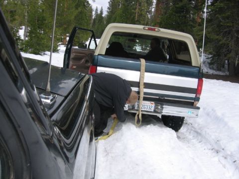 1989 Ford Bronco - A man secures a tow strap to a Ford Bronco stuck in a snowy area, with another vehicle nearby.
