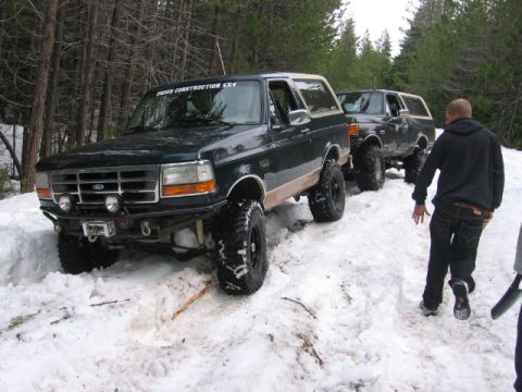 1989 Ford Bronco - Two lifted Ford Broncos navigate a snowy trail, one with custom text on its windshield.