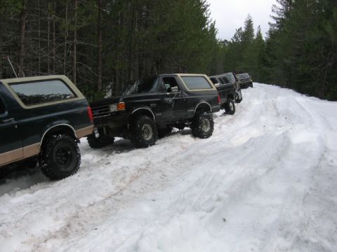 1989 Ford Bronco - Multiple Ford Bronco SUVs navigate a snowy forest road in a line.