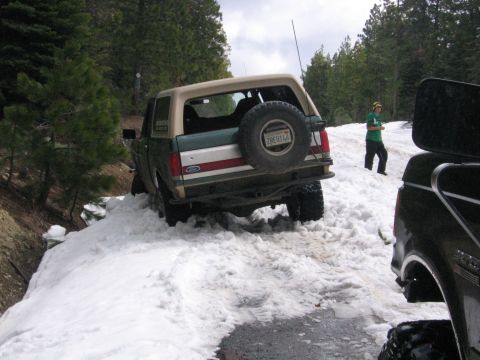 1989 Ford Bronco - A 1989 Ford Bronco navigates a snowy mountain road, its rear tire visible as it's partially stuck in the snow.