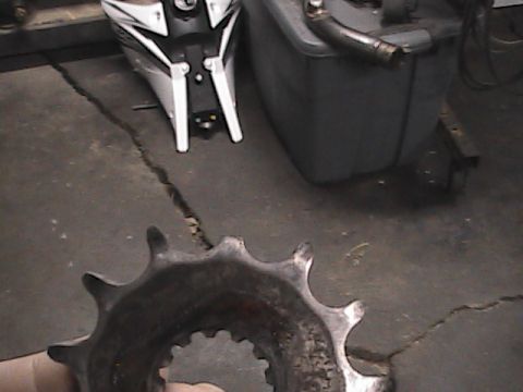 2007 Yamaha YZ450F - A worn motorcycle sprocket is shown in the foreground, with parts of a motorcycle and a fuel tank visible behind it the background.