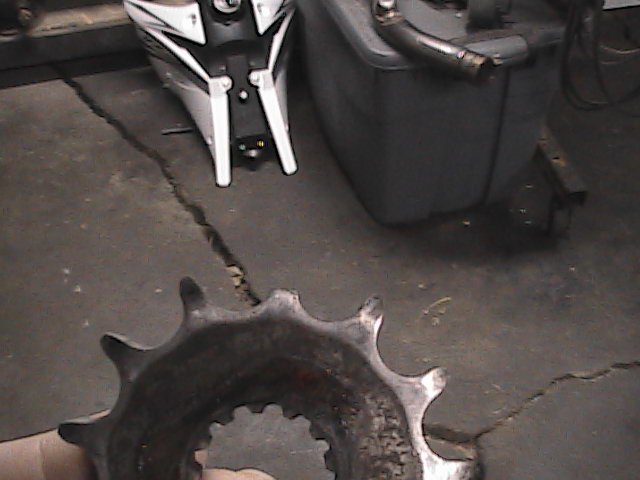 Close-up of a used motorcycle sprocket held in a hand, with a motorcycle body and a grey tank in the background