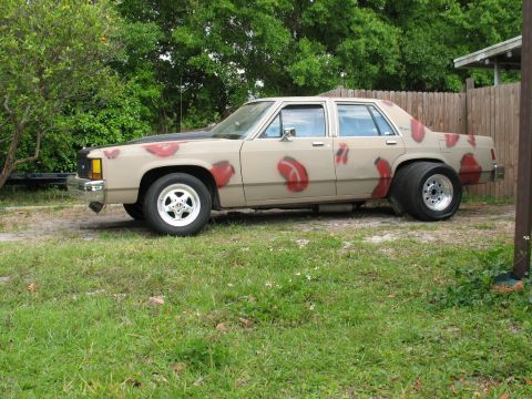 1985 Ford LTD Crown Victoria - A tan sedan with a black hood and red spray-painted shapes is parked on a grassy area.