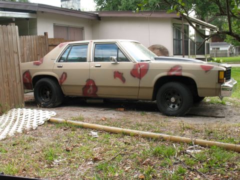 1985 Ford LTD Crown Victoria - A tan sedan with a black hood and red splotches on its body is parked in a grassy area.