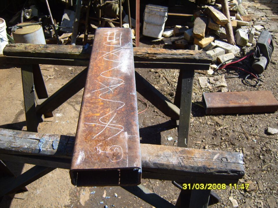 Rusty rectangular metal tube with chalk scribbles and markings, resting on sawhorses outdoors