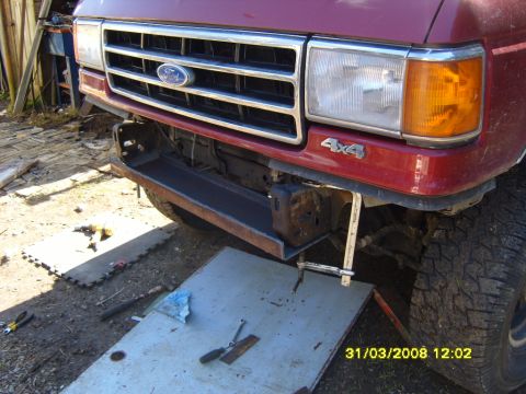 1989 Ford Bronco - Front end of a maroon/dark red 1980s Ford truck with the front bumper removed, showing the chrome grille with Ford oval, single rectangular…