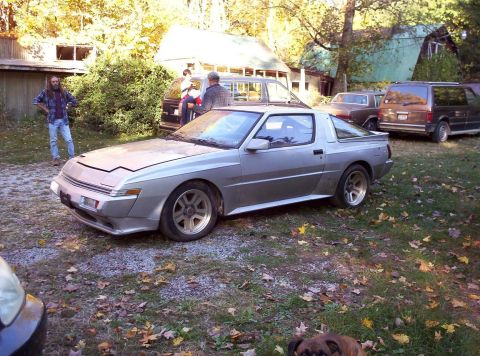 1995 Ford Probe - A silver Mitsubishi Starion sits in a yard with other vehicles and people nearby.